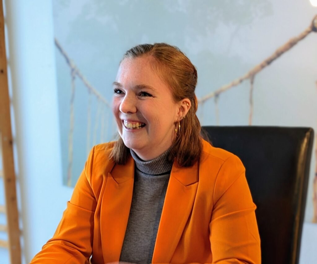 Frau rechts mit grauem Pullover und orangenem Blazer, lächelnd mit Zähnen, dunkelblonden Haaren, schaut nach links, mit verschwommenen Hintergrund