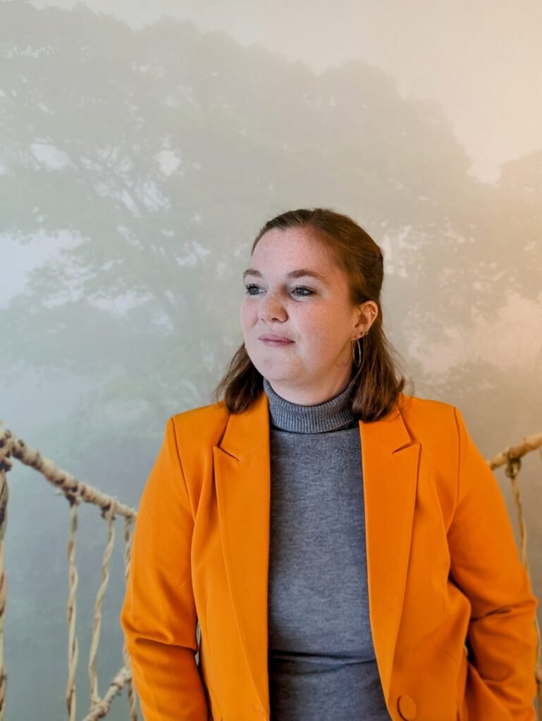 Frau mit grauem Pulli und orangenem Blazer und offenen, dunkelblonden Haaren schaut nach links vor verschwommenen, farbigem, Hintergrund