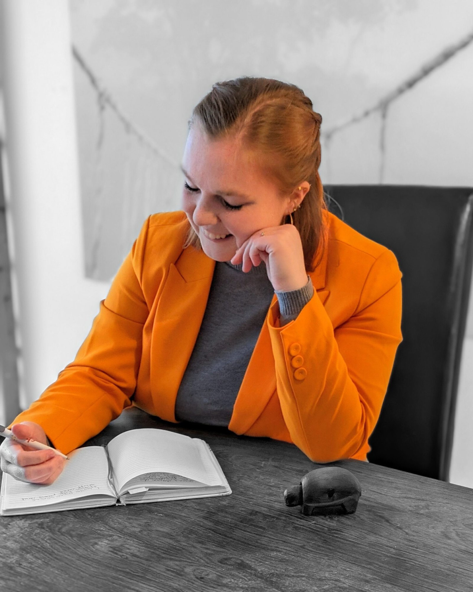 Frau mit grauem Pulli und orangenem Blazer hat den Kopf auf die Hand gestützt und den Ellbogen auf den Tisch an dem sie sitzt und schaut nach unten, sieht nachdenkend aus und hat dunkelblonde Haare vor schwarz-weißem Hintergrund
