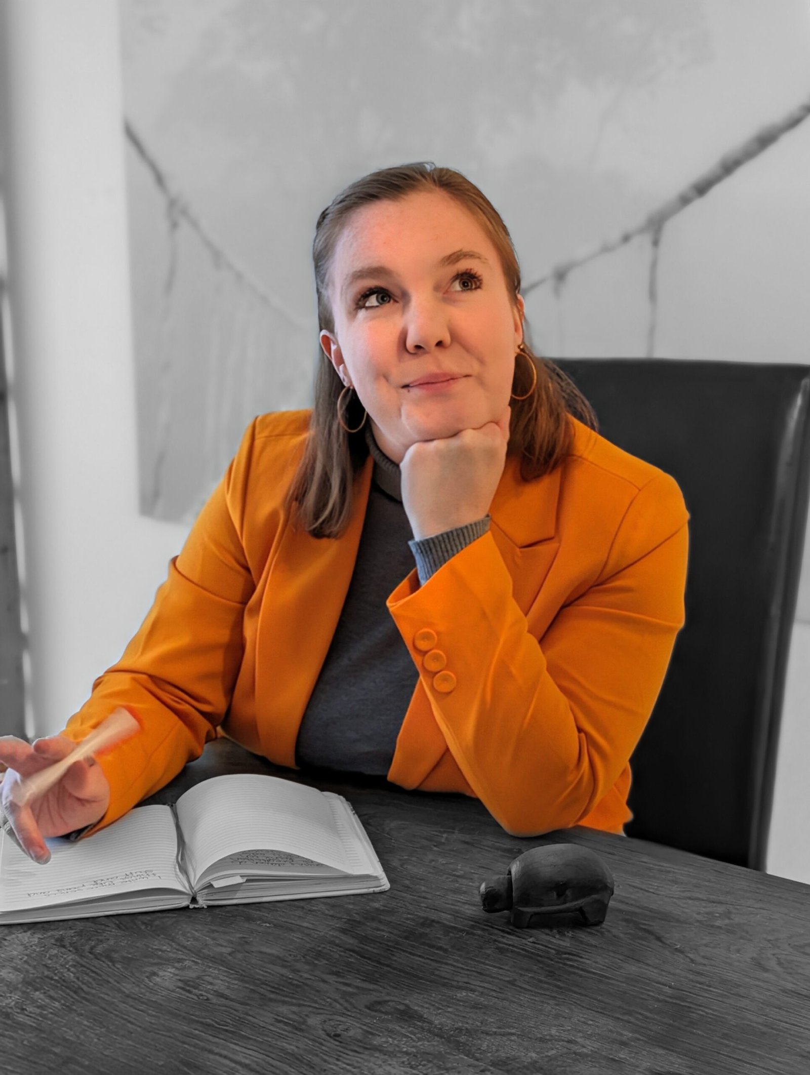 Frau mit grauem Pulli und orangenem Blazer hat den Kopf auf die Hand gestützt und den Ellbogen auf den Tisch an dem sie sitzt und schaut nach rechts, sieht nachdenkend aus und hat dunkelblonde Haare vor schwarz-weißem Hintergrund