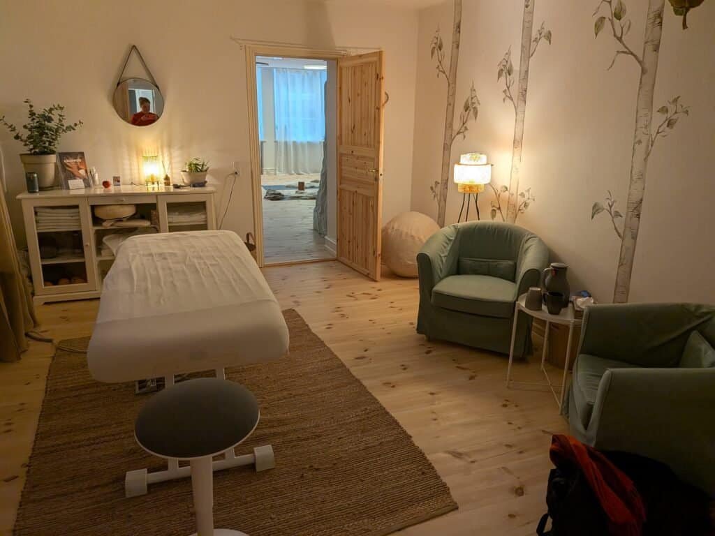 warm light, wooden floor, two green chairs, door in the background, massage table and mirror on the left
